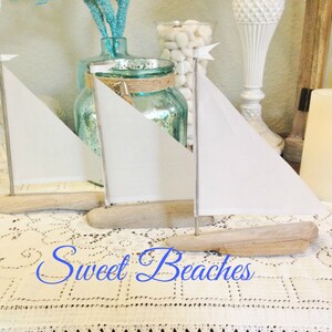 Puede incluir: Tres veleros blancos hechos de madera a la deriva con velas blancas. Los veleros est&aacute;n sentados sobre un mantel de encaje blanco. El texto "Sweet Beaches" est&aacute; escrito en cursiva azul sobre el mantel.