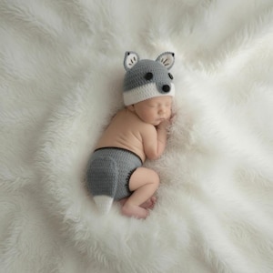 May include: A gray and white crocheted wolf hat with black button eyes and a nose, paired with matching gray and white crocheted diaper cover shorts featuring a small tail. The set is crafted with soft yarn and designed to resemble a wolf character.