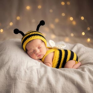 May include: A crocheted bee costume consisting of a yellow and black striped hat with two black antennae and a matching yellow and black striped romper with small white wings attached to the back, worn by a sleeping infant on a soft fabric.