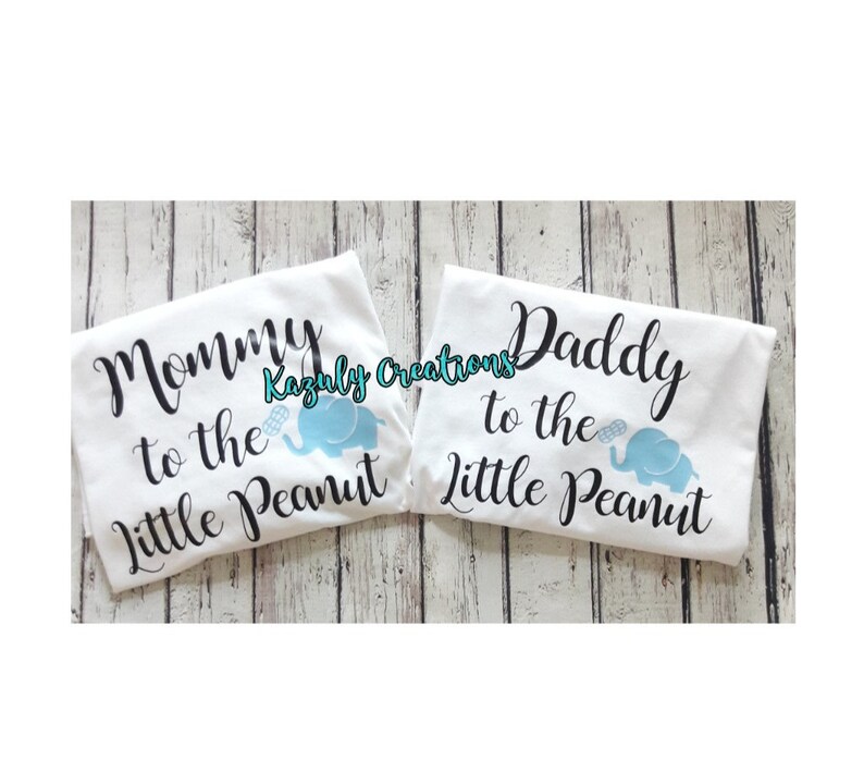 May include: Two white t-shirts with black text and a blue elephant graphic. The text on the left shirt reads "Mommy to the Little Peanut". The text on the right shirt reads "Daddy to the Little Peanut".