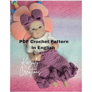 May include: A baby wearing a purple crochet outfit, including pants and a headband with a bow. The outfit is handmade and the text "PDF Crochet Pattern In English" is visible. The baby is lying on a pink and purple surface.