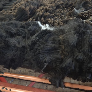 May include: A large collection of raw, dark brown and black sheep wool fleece spread across a red metal slatted drying rack. The wool appears thick, textured, and unwashed, with visible clumps of natural fibers arranged in a dense, layered pile.