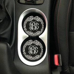 May include: Two black and white leopard print car coasters with the monogram "BC" in white. The coasters are in a car cup holder.