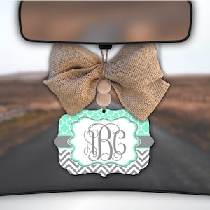 May include: A car air freshener with a gray and teal chevron pattern and a monogrammed initial "BC". The air freshener is hanging from the rearview mirror of a car. A burlap bow is attached to the top of the air freshener.