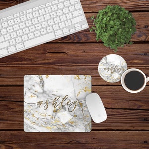 Peut inclure: Un tapis de souris en marbre blanc avec des accents dorés et le nom "ashley" écrit en cursive. Le tapis de souris est sur une surface en bois avec un clavier d'ordinateur blanc, une tasse à café blanche et un dessous-de-verre avec le même design que le tapis de souris.