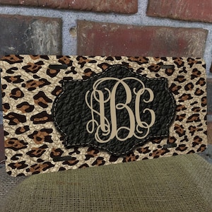 May include: A rectangular leopard print wallet with a black leather patch featuring a monogram. The wallet has a tan and brown spotted pattern. The monogram is in a light tan color.