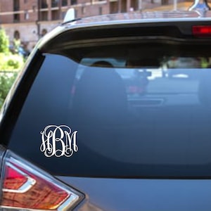 May include: A white monogram decal with the letters "LBM" in a script font on a gray car's rear window.