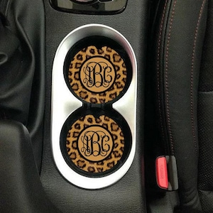 May include: Two leopard print car coasters with a monogram in a circle in the center. The coasters are in a car cup holder.