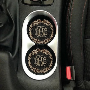 May include: Two black and brown leopard print car coasters with a white monogram "JBC" in the center. The coasters are in a car cup holder.
