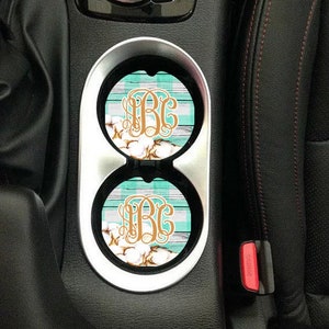 Könnte beinhalten: Zwei Auto-Untersetzer mit einem karierten Muster in Teal und Weiß und einem goldenen Monogramm "B.G." auf weißem Hintergrund. Die Untersetzer befinden sich in einem Getränkehalter im Auto.