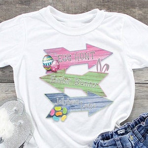 May include: White t-shirt with a graphic of three wooden arrows pointing in different directions. The arrows are decorated with Easter-themed designs and text, including "Egg Hunt", "Easter Bunny", and "Jelly Bean Lane".