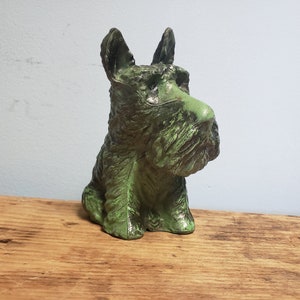 May include: A green ceramic figurine of a Scottish Terrier dog sitting with its head turned to the side. The figurine has a distressed finish and is detailed with textured fur.