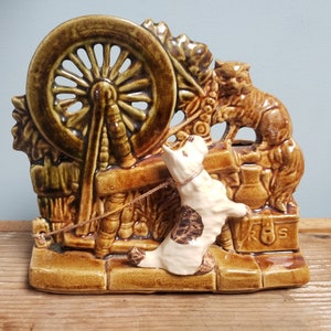 May include: A ceramic figurine of a cat and a dog with a spinning wheel. The cat is brown and white and the dog is white with brown spots. The spinning wheel is brown and green.
