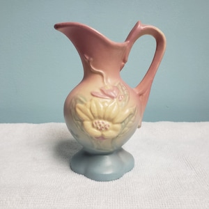 Könnte beinhalten: Eine antike Keramikkanne mit einem Blumendesign in Rosa-, Gelb- und Blautönen. Die Kanne hat einen breiten, runden Körper und einen zarten Henkel.