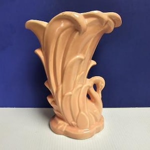 May include: A pink ceramic vase with a swan design. The vase is shaped like a fan and has a textured surface.