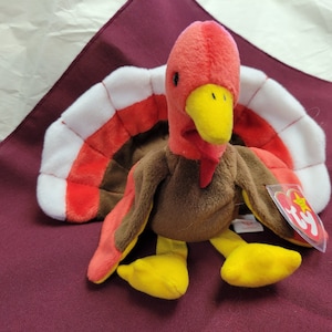 Puede incluir: Un pavo de peluche marrón y rojo con una cola en forma de abanico blanca y roja. El pavo tiene un pico y patas amarillas. El juguete tiene una etiqueta Ty adjunta.