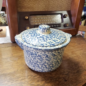 Puede incluir: Una fuente para horno vintage de gres moteado azul y blanco con tapa. El plato redondo tiene dos asas y una tapa a juego. El fondo presenta una radio de madera.