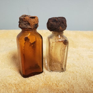 2 little Medicine Bottles -1 Brown Glass Bottle 3" tall x 1" x 3/4". 1 Clear Glass Bottle 3" tall x 1" x 1" with original corroded lids.