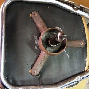 Może przedstawiać: Zbliżenie na czarną podstawę obrotową krzesła z metalową ramą i brązowym sk&oacute;rzanym siedziskiem. Podstawa ma metalową płytkę z nadrukowanym tekstem "FIXTURES MFG. CORP. 1541 Crystal St. K. C. 3, Mo. Corporation".