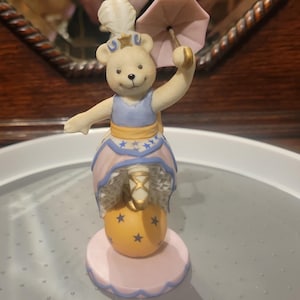 Puede incluir: Una figurita de porcelana de un oso de peluche vestido como un artista de circo, equilibrándose sobre una bola. El oso lleva un vestido azul y rosa con estrellas doradas y sostiene una sombrilla rosa.