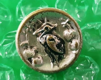May include: A silver and gold button with a detailed image of a bird with a long beak and a leafy branch with berries. The button has a raised edge and a smooth, shiny surface.