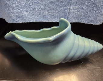 Könnte beinhalten: Eine hellblaue, keramik-Vase in Form einer Muschel. Die Vase hat eine breite Öffnung und einen langen, spiralförmigen Schwanz.