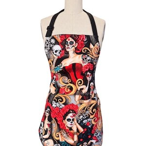 Puede incluir: Un delantal con un patrón repetitivo de figuras con maquillaje de calavera, rosas rojas y calaveras sobre un fondo negro. El delantal tiene tiras negras para el cuello con una hebilla ajustable y un dobladillo redondeado, sobre un maniquí.