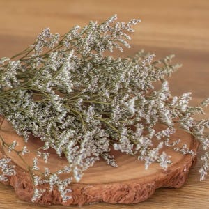 May include: A close-up of dried white flowers with green stems, arranged on a rustic, round wooden slice. The flowers have a delicate, textured appearance, and the wood slice has a natural, rough edge. The background is a warm, wooden surface.