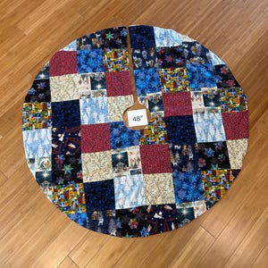 May include: A circular patchwork Christmas tree skirt featuring a variety of fabric squares in shades of blue, red, cream, and black. The design includes patterns of snowflakes, nativity scenes, and small village houses. The center has a 48 inch label.