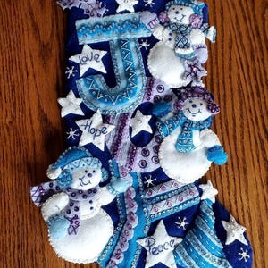 May include: Blue felt Christmas stocking with a white felt snowman design. The stocking is decorated with sequins, beads, and embroidered words "Love", "Hope", "Peace", and the letter "J".