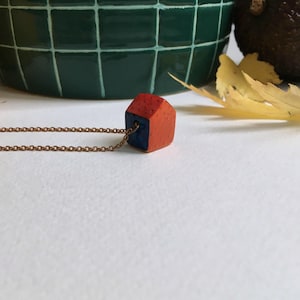 Op de afbeelding: Een rode en blauwe keramische huis hanger aan een gouden ketting ketting. Het huis is een geometrische vorm met een getextureerd oppervlak.
