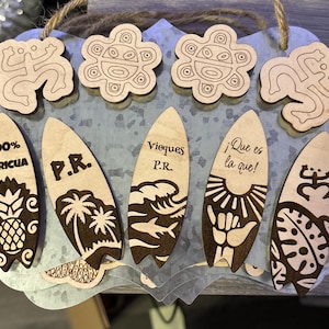 May include: Wooden decorative sign featuring surfboard-shaped ornaments with etched designs and text. The sign includes various symbols, text that reads "100% BORICUA", "P.R.", "Vieques P.R.", and "¡Que es la que!". The sign also has wooden cutouts.