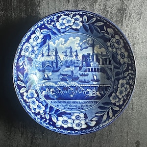 May include: Blue and white ceramic plate with a scene of ships and a building. The plate has a floral border and the text "Landing of Gen. Lafayette at Castle Garden, New York, 6th August 1824".