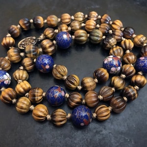 May include: A long necklace made of tiger's eye beads and blue and white enamel beads. The tiger's eye beads are brown and gold and have a ribbed texture. The enamel beads have a floral design.