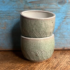 May include: Two stacked ceramic mugs with a green glaze and a textured surface. The mugs have a light brown rim.