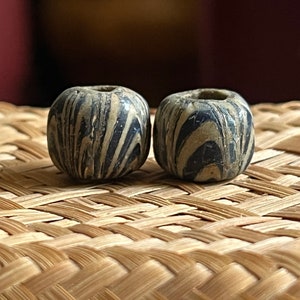 Può includere: Due perline rotonde in ceramica marmorizzata bianca e nera. Le perline hanno un motivo a spirale e sono su una superficie intrecciata.