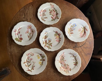 May include: Six white porcelain plates with scalloped edges and floral designs. Each plate features a different arrangement of pink, blue, and yellow flowers with green leaves.