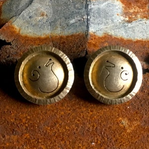 May include: A pair of gold-toned metal earrings with a stylized design of a bird or animal. The earrings are round and have a textured edge.