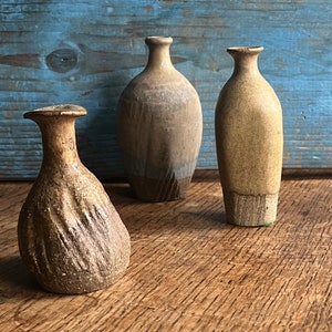 May include: Three ceramic vases in different shapes and sizes. The vases are all brown and have a rustic, handmade look. They are arranged on a wooden surface.