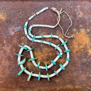 Puede incluir: Un collar artesanal con cuentas de concha blancas y detalles de color turquesa. El collar está ensartado en un cordón de color natural y tiene un diseño único y orgánico. El collar se muestra sobre una superficie marrón oxidada.