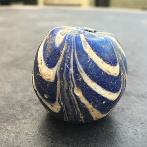 Puede incluir: Una cuenta de vidrio azul y blanco con un patrón de espiral. La cuenta es redonda y tiene un agujero en el centro.