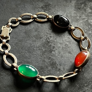 May include: A silver chain bracelet with three oval-shaped gemstones: a black stone, a green stone, and a red stone.