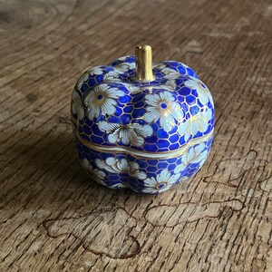 Könnte beinhalten: Eine kleine, blaue und weiße Keramik-Schmuckschatulle in Form eines Kürbisses mit goldener Oberseite. Die Schachtel ist mit weißen Blumen und einem Wabenmuster verziert.