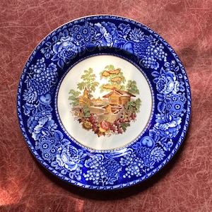 May include: A round, blue and white decorative plate. The plate features a detailed floral and fruit border. The center of the plate has a landscape scene with buildings and trees. The plate has a gold rim.
