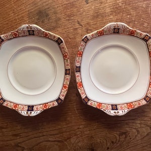 May include: Two square ceramic plates with a white background and a decorative border in shades of blue, orange, and gold. The plates have scalloped edges and a raised rim.