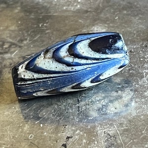 May include: A blue and white glass bead with a swirling pattern. The bead is oval shaped and has a glossy finish.