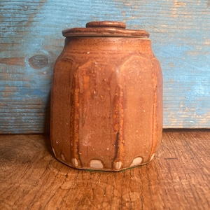 Puede incluir: Un tarro de cerámica marrón con tapa. El tarro tiene una superficie texturizada y una forma ligeramente redondeada. La tapa también es marrón y tiene una superficie lisa.