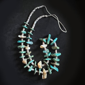 May include: A turquoise and off-white beaded necklace with a silver-colored cord. The necklace is composed of irregularly shaped turquoise and off-white beads, strung together with a silver-colored cord. The necklace is displayed against a black background.
