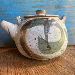 Könnte beinhalten: Eine weiße Teekanne aus Keramik mit einem braunen Deckel und braunen, grünen und blauen abstrakten Mustern. Die Teekanne hat einen runden Körper und einen Ausguss an der Seite.