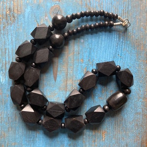May include: A black wooden necklace with large, faceted beads. The necklace is made of two strands of beads, with one strand being smaller than the other. The beads are all black and have a smooth, polished finish.